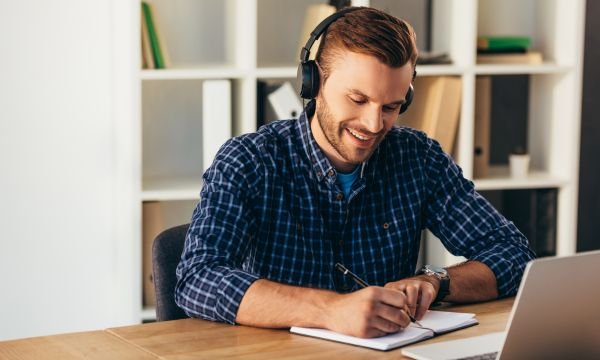 portrait of smiling white man in headphones making notes while taking part in webinar at tabletop with laptop in office