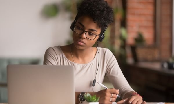 Focused mixed race woman wearing headphones watching webinar writing notes