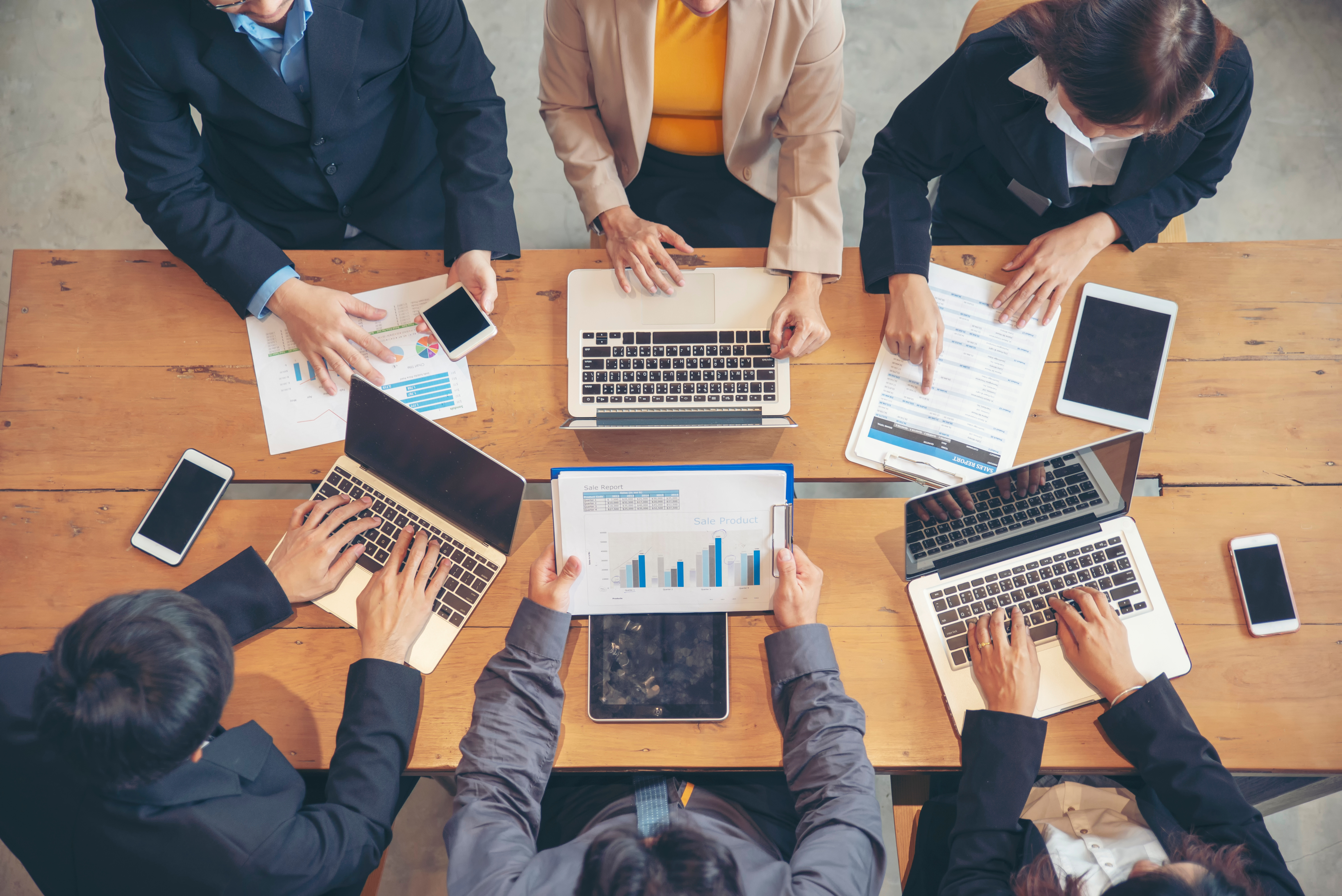 Top-down view of a business team analyzing sales charts and data dashboards on laptops and tablets around a wooden table.