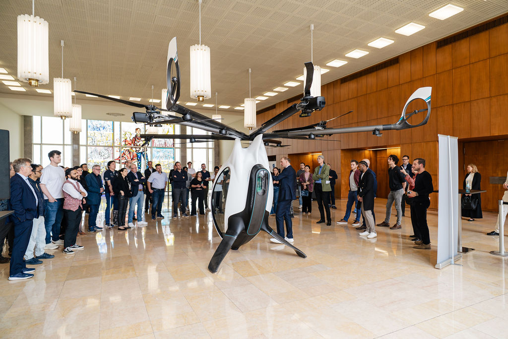 A group of people gathers around a prototype AirScooter flying vehicle inside ESMT Berlin’s foyer during a live demonstration.
