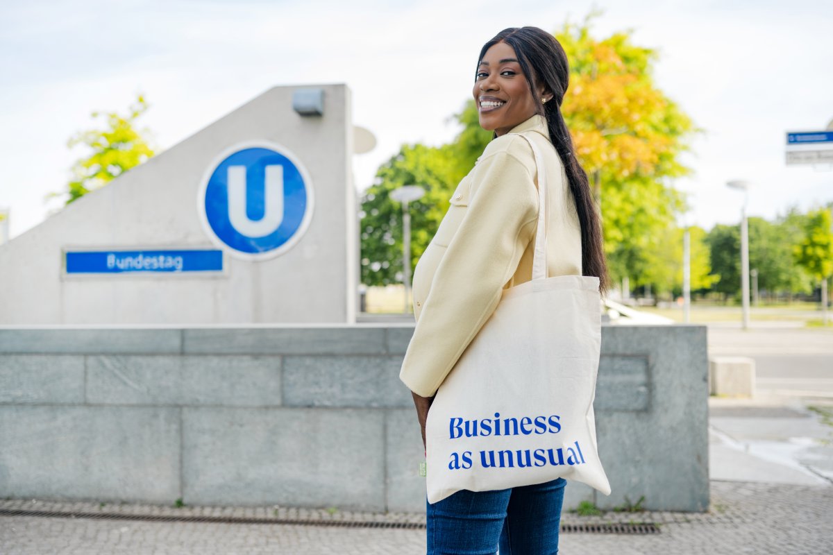 ESMT Berlin Full-time MBA student Gloria Motopu standing in front of a Berlin U-bahn station. She has a cream coloured blazer, long black hair, and an ESMT-branded tote