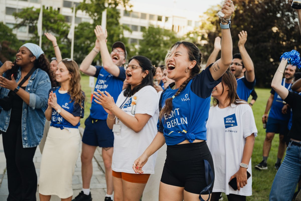 ESMT Berlin Full-time MBA students Ting Han, Roshni Dutta, Tammy Sokari, Jasmin Schubach and Christopher Gerbino cheering during the school Olympics. They are all wearing ESMT-brand tee shirts and are standing in the campus garden on a sunny evening.