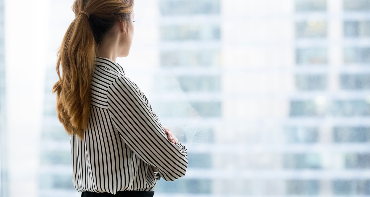 a solo businesswoman lost in thought looks through a glass window at the city below