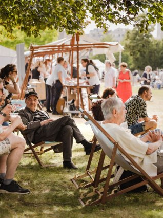 People relaxing in wooden deck chairs under a shady tree at a sunlit outdoor festival with tents and stalls in the background.