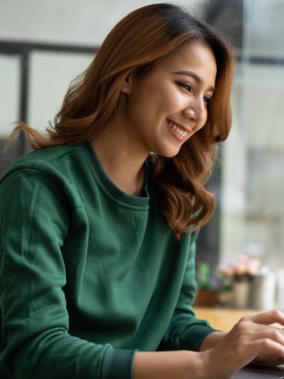 Woman in her early thirties sitting in a cafe and typing on her laptop. She has long brown hair and a silky emerald shirt.