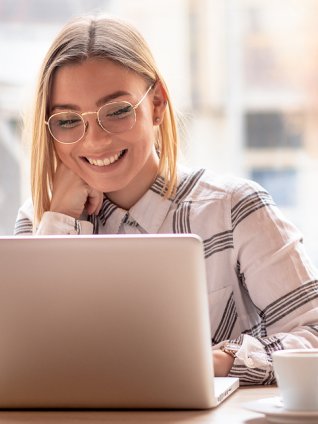 Woman in her early twenties smiling at the webinar on her laptop. She has glasses, chin-length blond hair, and is sitting in an office in the sunshine
