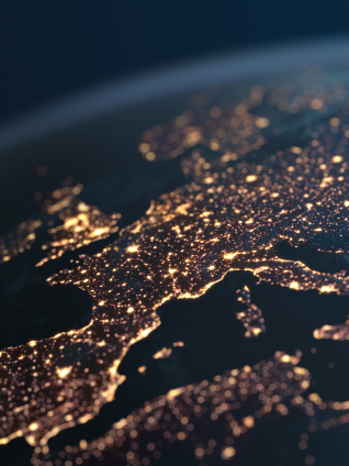 Nighttime satellite-style view of Europe and the Mediterranean region, with dense clusters of glowing city lights outlining coastlines and major urban areas against a dark, cloud-streaked background.