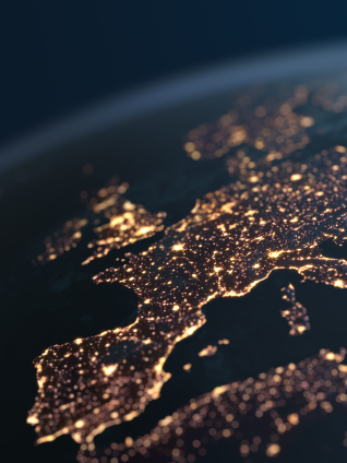 Nighttime satellite-style view of Europe and the Mediterranean region, with dense clusters of glowing city lights outlining coastlines and major urban areas against a dark, cloud-streaked background.