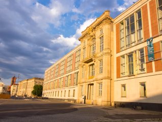 he grand exterior facade of the ESMT Berlin building at Schlossplatz, showcasing a historic, classical stone portal integrated into a modernist architectural structure under a partly cloudy sky in the warm evening light.