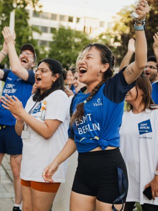 ESMT Berlin Full-time MBA students Ting Han, Roshni Dutta, Tammy Sokari, Jasmin Schubach and Christopher Gerbino cheering during the school Olympics. They are all wearing ESMT-brand tee shirts and are standing in the campus garden on a sunny evening.