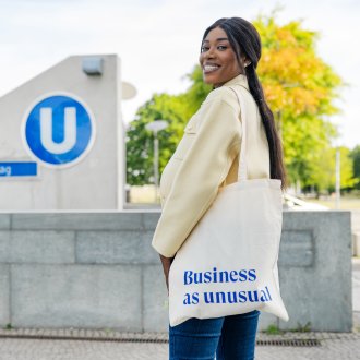 ESMT Berlin Full-time MBA student Gloria Motopu standing in front of a Berlin U-bahn station. She has a cream coloured blazer, long black hair, and an ESMT-branded tote