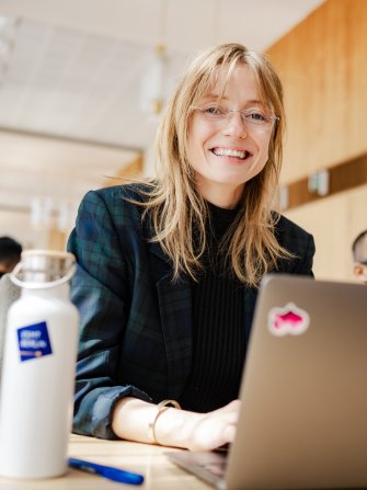 Picture of Johanne Björklund Larsen, an ESMT Berlin Full-time MBA student, working on her laptop and smiling at the camera. She has an ESMT branded waterbottle and is sitting at a desk.