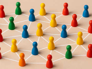 A photograph captures a vibrant network of multicolored wooden meeples, connected by crisp white lines against a neutral beige background. 