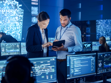 Two people working in a room filled with multiple monitors and computers, focused on their tasks.