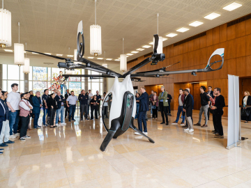 A group of people gathers around a prototype AirScooter flying vehicle inside ESMT Berlin’s foyer during a live demonstration.