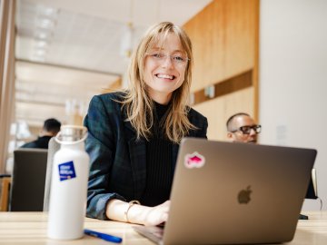 Picture of Johanne Björklund Larsen, an ESMT Berlin Full-time MBA student, working on her laptop and smiling at the camera. She has an ESMT branded waterbottle and is sitting at a desk.