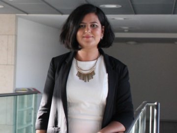 A woman with short dark hair stands indoors by a railing, wearing a black blazer over a white dress and a statement necklace, looking at the camera with a slight smile.