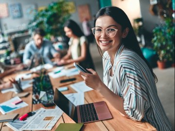 Woman in her twenties smiling and holding a smart phone and looking at a laptop. Her friends are at the other end of the table while she has a meeting online.