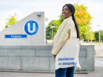ESMT Berlin Full-time MBA student Gloria Motopu standing in front of a Berlin U-bahn station. She has a cream coloured blazer, long black hair, and an ESMT-branded tote