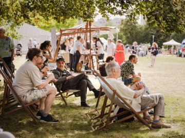 People relaxing in wooden deck chairs under a shady tree at a sunlit outdoor festival with tents and stalls in the background.