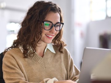 Woman in her early twenties watching a webinar and smiling. She has dark curly hair and glasses