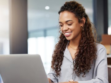 New graduate watching a webinar in her office. She is dressed professionally, and has long, slightly curly hair.