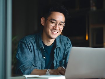 Man in his thirties watching a webinar on his home laptop. He is wearing glasses and smiling with interest
