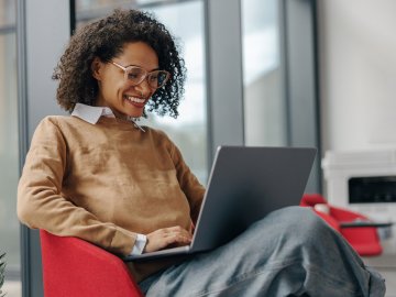 Woman with very curly hair sitting in an office armchair and watching a webinar on her laptop