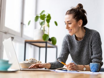 Woman in a grey sweater watching an online webinar while having breakfast at home