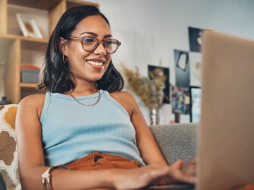 Woman in her late thirties with glasses and a big smile. She is sitting on her sofa and watching a webinar on her laptop from the comfort of home