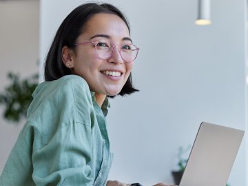 Woman in her twenties looking up from her laptop and smiling. She has pink glasses and a mint green shirt