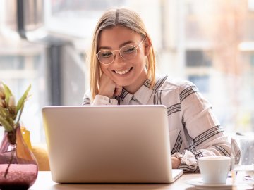 Woman in her early twenties smiling at the webinar on her laptop. She has glasses, chin-length blond hair, and is sitting in an office in the sunshine