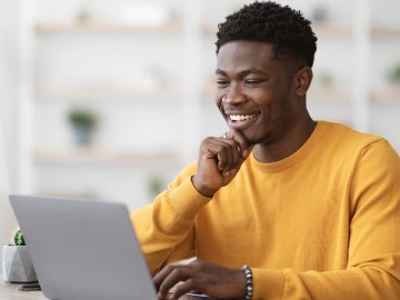 Man in his twenties in a bright yellow sweater. He has very curly black hair, and is looking at the webinar on his laptop in a happy but intrigued way