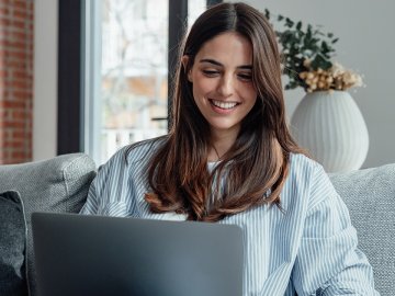 Woman in her twenties sitting on her sofa in a blue and white striped shirt. She is watching a webinar on her laptop