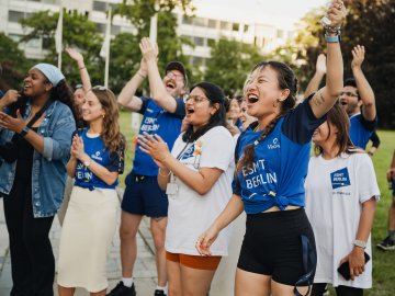ESMT Berlin Full-time MBA students Ting Han, Roshni Dutta, Tammy Sokari, Jasmin Schubach and Christopher Gerbino cheering during the school Olympics. They are all wearing ESMT-brand tee shirts and are standing in the campus garden on a sunny evening.