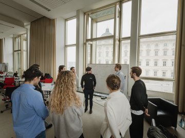 ESMT students taking part in a business-development bootcamp on campus. They are standing at a white board discussing a concept. The Humboldt forum is visible through the window behind them.