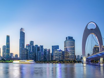 A panoramic view of the Guangzhou city skyline, featuring modern skyscrapers along the Pearl River with Canton Tower visible in the distance under a blue sky.
