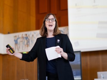 Beth Sibly gives talk holding notes and remote in lecture hall, slide projected behind.