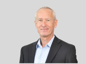 Professional headshot of smiling gray-haired man in suit against gray background.
