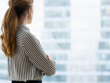 a solo businesswoman lost in thought looks through a glass window at the city below