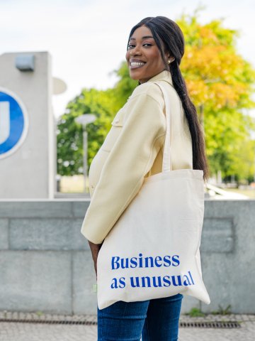 ESMT Berlin Full-time MBA student Gloria Motopu standing in front of a Berlin U-bahn station. She has a cream coloured blazer, long black hair, and an ESMT-branded tote