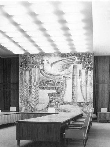 A black-and-white historical photograph of a large, formal conference room. It features a long U-shaped wooden table with modern chairs, wood-paneled walls, and a large mosaic mural at the far end depicting a dove and stalks of grain. The ceiling is covered with a grid of square light panels.