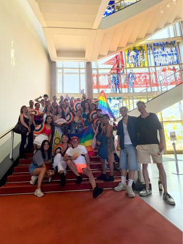 Students from the ESMT Berlin Pride Impact Club posing on the main campus staircase. They are carrying rainbow fans and look joyful.