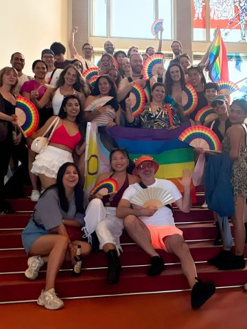 Students from the ESMT Berlin Pride Impact Club posing on the main campus staircase. They are carrying rainbow fans and look joyful.