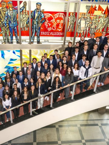 The ESMT Berlin Master in Global Management cohort, Class of 2026. They are standing on the main campus staircase in front of the historic, three-story stained glass window