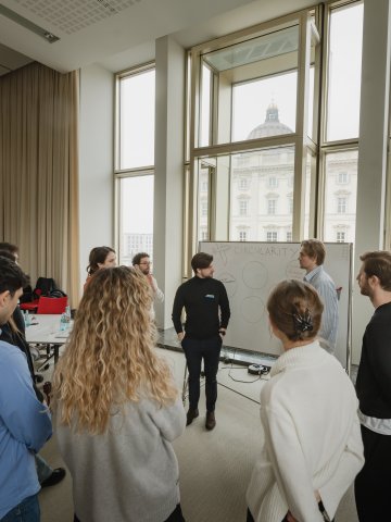 ESMT students taking part in a business-development bootcamp on campus. They are standing at a white board discussing a concept. The Humboldt forum is visible through the window behind them.
