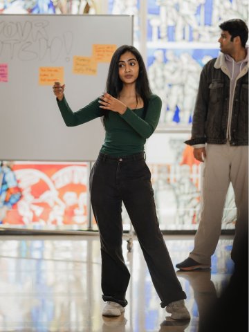 An ESMT Berlin master's student demonstrating a concept by moving her arms. She is in a dark green shirt and is standing in front of a whiteboard on the ESMT campus