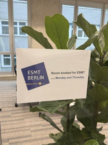 Printed ESMT Berlin room booking sign taped to glass, plant and windows behind