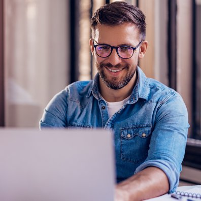 Man in his thirties watching a webinar on his laptop. He is wearing a blue button down shirt and glasses and is looking pleased