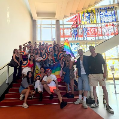 Students from the ESMT Berlin Pride Impact Club posing on the main campus staircase. They are carrying rainbow fans and look joyful.